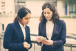 © Mangostar - Focused businesswomen using smartphone. Female colleagues in formal wear standing on street and using cell phone together. Business and technology concept
