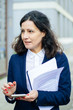 © Mangostar - Professional businesswoman with smartphone and papers. Focused middle aged businesswoman holding cell phone and documents while standing and looking aside on street. Business concept