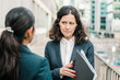 © Mangostar - Serious businesswomen discussing work. Professional female colleagues in formal wear standing on urban city street and discussing papers. Cooperation concept