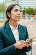 © Mangostar - Pensive businesswoman using smartphone. Thoughtful young businesswoman in formal wear holding cell phone and looking aside on street. Communication concept