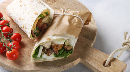 Caesar wrap. Chicken, lettuce, egg, sauce and salad in pita bread on wooden board on the white table. Rolls in parchment, cherry tomatoes. Shawarma horizontal. Background image, copy space