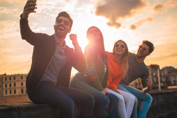 Group of happy young influencers people taking a selfie sitting on a bench outdoors in the sunset. Real backlight flare. Leisure activities lifestyle concept.