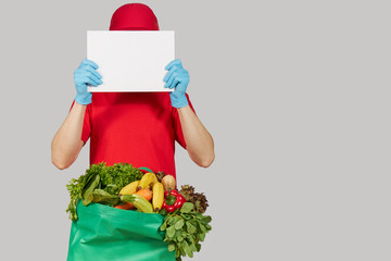 Online shopping concept. Courier in red uniform, protective mask and gloves with a grocery box with fresh fruits and vegetables holds a White banner for text. Home delivery food during quarantine 