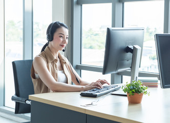 Attractive and smile young asian customer servicer woman wearing headset in modern creative meeting working office look at computer monitor,work from and social distancing concept.