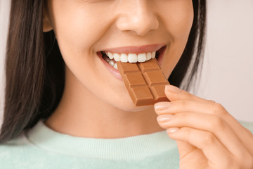 Poster - Beautiful young woman eating tasty chocolate on light background, closeup