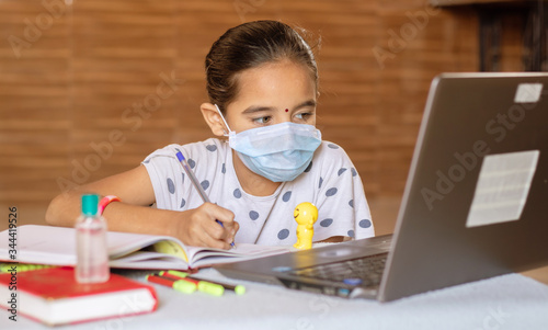 Concept of homeschooling and e-learning, young girl with medical mask writing by looking into laptop during covid-19 or coronavirus pandemic lock down.