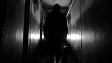 Silhouette Of Unrecognizable Man In Black Jacket And Carrying A Bag Confidently Walking Away From The Camera Along Hallway In Old Apartment Building Long Dark Hallway. Criminal Concept