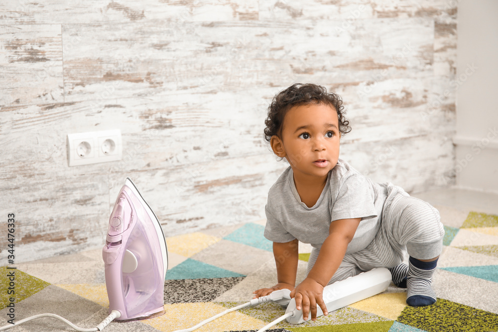 Little African-American baby playing with iron at home. Child in danger