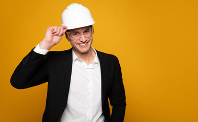 Let me introduce myself. Young handsome man in a suit is tipping his hard hat while looking in the camera and smiling.