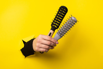 female hand holds two hairdresser combs from a torn hole in yellow paper