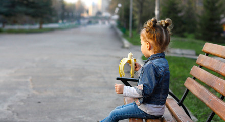 little girl in denim clothes sits on a wooden bench in the park and eats a banana.