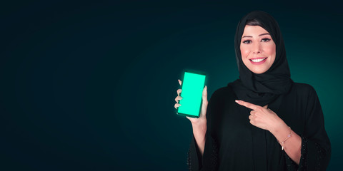 Cheerful cute Arab woman pointing finger on smartphone screen isolated on a black background. Looking at camera, technology concept.