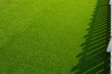 Sunlight and shadow of white wooden fence on surface of green artificial turf in front yard of home 