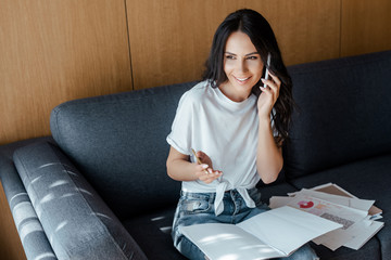 happy woman talking on smartphone and working with business documents on sofa during self isolation