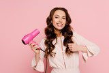 cheerful young woman holding hair dryer and showing thumb up isolated on pink