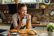 Leinwandbild Motiv Happy woman eating slice of bread while using smart phone in the kitchen.