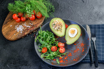 Wall Mural - Ketogenic diet food dinner of fresh avocado, tomatoes cherry, arugula and fresh green herbs on the black background. Avocado with filling of egg and tuna with sauce. Healthy keto diet food.