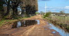 A Rural Country Road Full Of Chuckholes, Potholes, And Disrepair Holds Water After An Overnight Rain. It Could Use Some Repair Work And Loads Of Gravel.