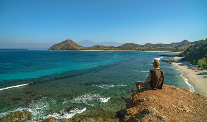 Wall Mural - Blond man sitting and enjoying the views to undiscovered beach, hills and Rinjani Volcano seen in Sumbawa island