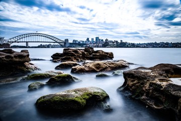 Long exposure on a partly cloudy afternoon on Sydney Harbour with nice rocks in the foreground the soft waves crashing on the shore and the beautiful harbour foreshore as a backdrop