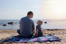 Dad And Son Are Sitting On The Beach And Chatting. A Boy With A Father On The Background Of The Sea And Sunset Cuddling. The Concept Of A Happy Childhood And Hanging Out With A Parent