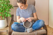 a young girl is sitting at home in quarantine in a chair and crocheting a basket made of gray knitted cotton yarn close up in blue jeans and a striped t shirt