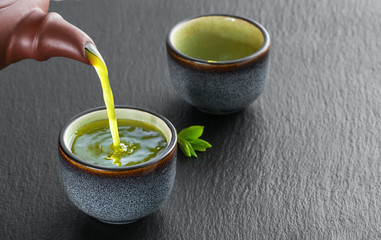 Hot green tea is poured from the teapot into the blue bowl, darck stjne table. Tea leaves next to the cup. Close-up, tea ceremony, minimalism, copy space for text.