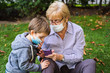 © progat - Grandmother and little kid playing with a smartphone in the backyard while wearing protective masks due to coronavirus outbreak
