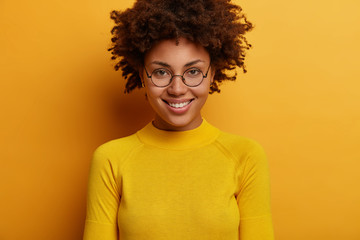 Wall Mural - Portrait of lovely woman smiles gently, has curly hair, wears round transparent glasses and yellow jumper, looks directly at camera, listens pleasant news, poses indoor. Human face expressions