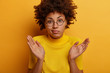 © wayhome.studio  - Confused dark skinned female spreads palms and shruggs shoulders, feels indecisive, asks question, doesnt understand what happening, looks through transparent glasses, isolated on yellow background