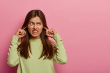 Resentful Annoyed Woman Plugs Ears, Irritated With Loud Noise, Avoids Annoying Sound, Dressed In Casual Green Jumper, Clenches Teeth, Frowns Face, Isolated On Pink Background, Blank Space Aside
