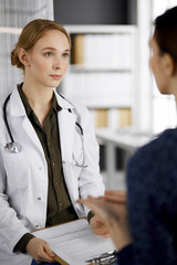 Friendly smiling female doctor and patient woman discussing current health examination while sitting in clinic. Medicine and healthcare concept