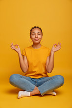 Peaceful Calm Dark Skinned Female Model Sits Crossed Legs On Floor, Practices Yoga And Tries To Relax, Breathes Deeply, Closes Eyes, Reaches Nirvana, Holds Hands Sideways, Releases Stress After Work