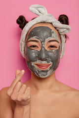 Wall Mural - Natural Asian woman has healthy skin, makes mini heart gesture, stands with bare shoulders, applies beauty mask for rejuvenation, wears headband, isolated on pink background. Face care routine