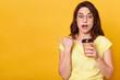 © sementsova321 - Closeup portrait of astonished woman posing against yellow studio background, looks surprised, being shocked, holding takeaway coffee, pointing aside, keeps mouth opened. Copy space for advertisement.