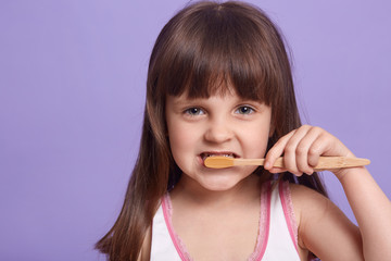 Wall Mural - Close up portrait of caring sweet kid looking directly at camera, brushing teeth, keep clean, having straight hair, wearing shirt, posing isolated over lilac background in studio. Hygiene concept.