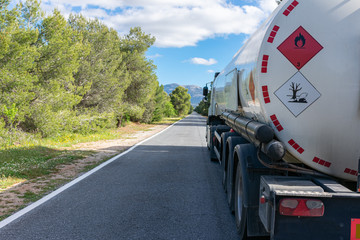 Wall Mural - Hazard labels for flammable and polluting liquid on a fuel tank truck driving on a narrow, straight road lined with trees.