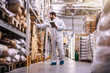 © Dusan Petkovic - Full length of young employee in sterile protective uniform checking on products while standing in warehouse full of food products in boxes.