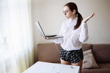 Home office dress code. A girl in a strict white shirt is talking over a video link. On her feet, she is wearing homemade pajama shorts. The atmosphere is homely and cozy.