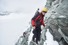 Full Length Of Male Mountaineer In Sunglasses Using Fixed Rope To Climb Winter Mountain. Alpinist In Safety Helmet Standing On Rock Covered With Snow. Concept Of Mountaineering, Alpine Rock Climbing.