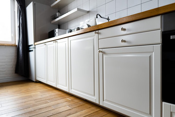 Appliances and furniture: bright white cabinets with wooden countertop, electric cooker, induction hob, faucet, sink and dish rack against hardwood floor and light tile in modern scandinavian kitchen