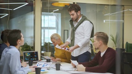 Wall Mural - Through glass wall tracking shot of business team planning work sitting at table during meeting in boardroom. Creative man standing and sharing ideas