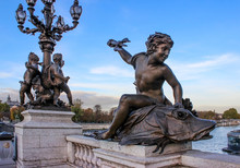 Sculpture On The Bridge. The Boy Is Sitting On A Fish, Holding A Trident. A Lantern In The Distance. France. Paris.