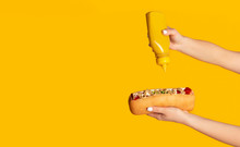 Unrecognizable Woman Squeezing Mustard Onto Hot Dog Against Orange Background, Blank Space. Panorama