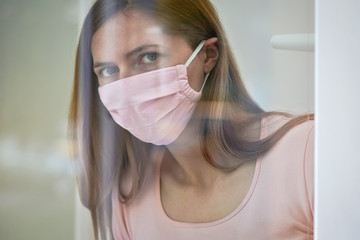 Wall Mural - Young woman in pink cotton face virus mask standing behind window glass pane, touching it with hand, looking sad. Quarantine or stay at home to be safe during coronavirus covid-19 outbreak concept