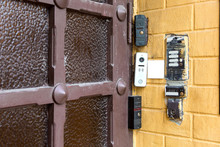Several Types Of Different Intercoms And Electronic Locks On A Chip Or Card On A Yellow Brick Wall On Outdoor Against A Wooden Door With Glass Inserts, Closeup Nobody.