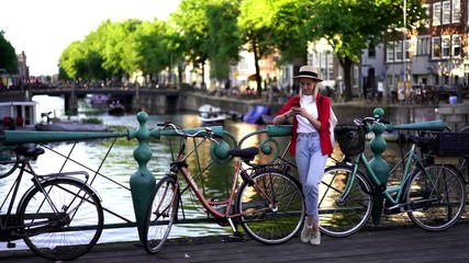 Wall Mural - Happy female tourist searching sightseeing in Amsterdam city using app on modern smartphone in roaming, positive hipster girl standing near famous canal bridge while browsing interior. Slow motion
