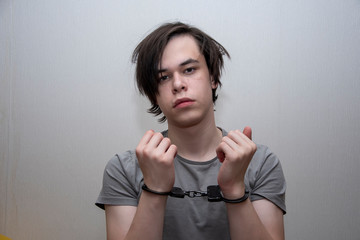 Portrait of a teenager in handcuffs on a gray background, medium plan. Juvenile delinquent, criminal liability of minors. Members of youth criminal groups and gangs.