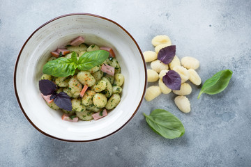 Wall Mural - Gnocchi with green basil pesto and sliced bacon in a white bowl, above view on a light-blue stone background, studio shot