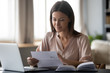 © fizkes - Young woman holding paper, reading good news in letter, sitting at desk with laptop at home, beautiful female received correspondence, bank statement or notification, pleasant message